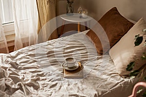 A cup of hot cappuccino, a book, glasses on the bed. Breakfast. Cozy house. Decor, details