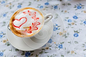 A Cup of hot cappuccino with a pattern of a red heart on the table with a tablecloth in a flower in a coffee shop. Close-up and bl