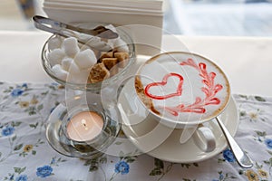 A Cup of hot cappuccino with a pattern of a red heart on the table with a tablecloth in a flower in a coffee shop. Near sugar bowl