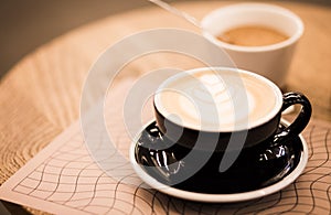 Cup of hot cappucino in a blak cup with coffee art on wooden table background. Hippster style