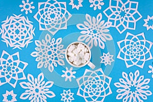 Cup of hot chocolate with marshmallow and paper snowflakes on blue background. Top view. Christmas decoration