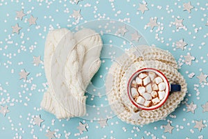 Cup of hot cocoa or chocolate with marshmallow and knitted mittens on blue winter background top view. Flat lay.