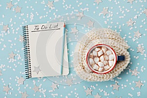 Cup of hot cocoa or chocolate with marshmallow and notebook with to do list on blue confetti background top view. Christmas plan.