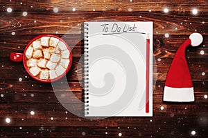 Cup of hot cocoa or chocolate with marshmallow, santa hat and notebook with to do list on table from above, christmas planning.