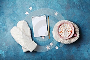 Cup of hot cocoa decorated knitted scarf with marshmallow, mittens and clean notebook on turquoise table top view. Flat lay style.