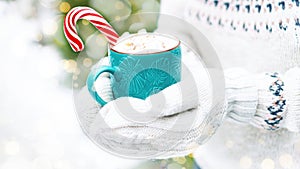 A cup of hot cocoa with marshmallows and candy cane in hands with knitted mittens on a snowy background with bokeh. Hot drinks for