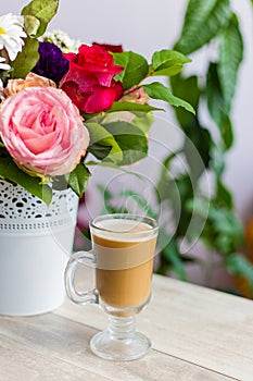 Cup of hot coffee with milk stands near the window and a bright colored bouquet of flowers