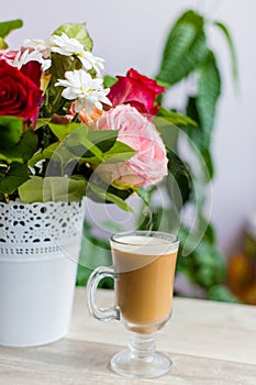 Cup of hot coffee with milk stands near the window and a bright colored bouquet of flowers