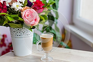 Cup of hot coffee with milk stands near the window and a bright colored bouquet of flowers