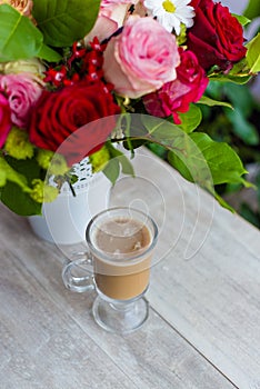 Cup of hot coffee with milk stands near the window and a bright colored bouquet of flowers