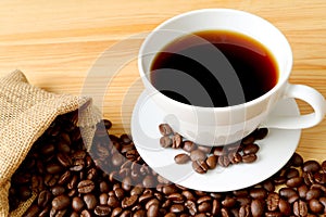 Cup of hot coffee with roasted coffee beans scattered from burlap bag on wooden table