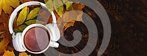 A Cup of hot coffee or tea with autumn leaves and headphones on a brown wooden background. banner