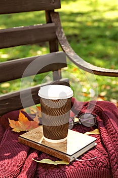 Cup of hot coffee with warm sweater and autumn leaves on wooden bench in park