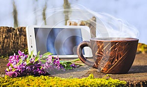 A cup of hot coffee in the woods on a stump next to the phone and spring flowers. Active rest in nature_