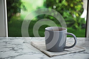 Cup of hot drink and napkin on marble windowsill against glass with rain drops