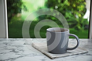 Cup of hot drink and napkin on marble windowsill against glass with rain drops photo