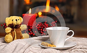 Cup of hot drink teddy bear candle in red Christmas decoration on cozy knitted plaid in front of warm fireplace. Holiday Christmas