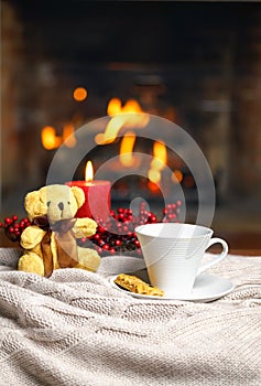 Cup of hot drink teddy bear candle in red Christmas decoration on cozy knitted plaid in front of warm fireplace. Holiday Christmas