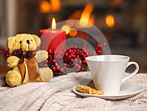 Cup of hot drink teddy bear candle in red Christmas decoration on cozy knitted plaid in front of warm fireplace. Holiday Christmas