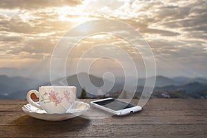 A cup of hot espresso coffee mugs placed with smartphone on a wooden floor with morning fog and moutains with sunlight background,