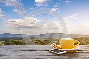 A cup of hot espresso coffee mugs placed with smartphone on a wooden floor with morning fog and moutains with sunlight background,