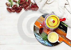 Cup of hot herbal tea, bright colored teapot on wooden tray blackboard, knitted plaid or sweater red chrysanthemum flowers on ligh