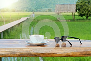 cup of hot milk coffee with sunglasses on wooden table in the morning at green rice field nature background with copy space