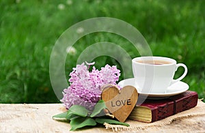 A cup of hot tea, a book and lilacs against a background of green grass. Romantic concept. Wooden card with a heart.