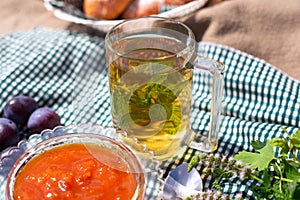 Cup of hot tea with mint in a transparent glass, next to a vase with berry jam in the fresh air. Seasonal still life