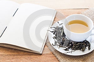 Cup of hot tea on sackcloth with dried tea leaves and handmade book on wooden table with copy space