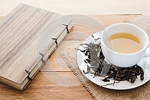 Cup of hot tea on sackcloth with dried tea leaves and handmade book on wooden table with copy space from high angle view.