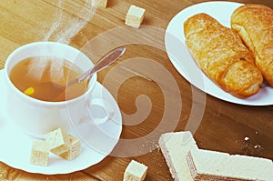 Cup of hot tea with steam and a croissant, wafer and toast on a old wooden table