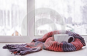 A cup of hot tea warmed in the knitted scarf is on the windowsill against the winter window