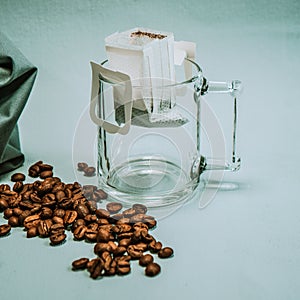Ground coffee, a bag for coffee drops and a transparent mug on the table. Drop coffee on a gray background with coffee beans.