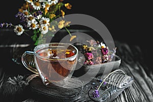 Cup of medicinal herbal tea with chamomile flowers and a bouquet of wild flowers on a rustic wooden background. Rustic still life