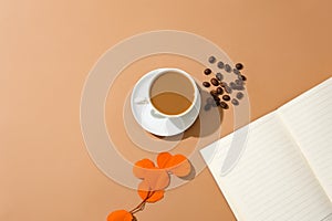 A cup of milky coffee, coffee beans, a notebook and a branch of autumn leaves are displayed on a brown background. Coffee beans