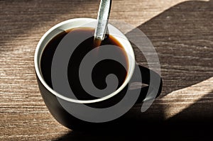 A cup of morning black coffee without milk with a spoon in a black cup on a wooden table. Shadows from the morning sun