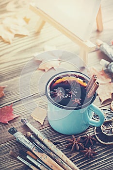 Cup of mulled wine, artist paintbrushes, paints, artistic canvas on easel and autumn leaves.