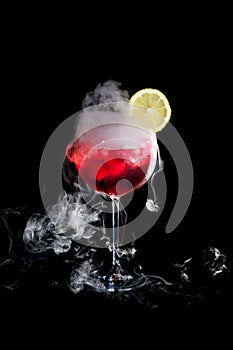 A cup of red cold cocktail with dry ice smoke and a lemon slice