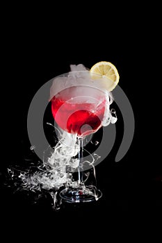 A cup of red cold cocktail with dry ice smoke and a lemon slice