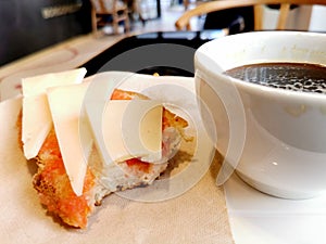 A cup of remaining black coffee with a bitten ham cheese sandwich on a cafe table