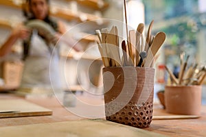 Cup with set of dirty art and craft sculpting tools on wooden table in pottery workshop, used to work clay and ceramic