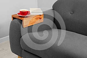 Cup of tea and book on sofa with wooden armrest table in room. Interior element