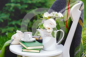 Cup of tea on books, flowers white wild rose in vase teapot, warm plaid on white chair outside in summer garden. Romantic provence