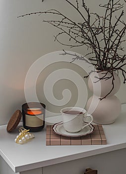 A cup of tea, a burning candle, a bouquet of dried branches in a ceramic vase on a white chest of drawers