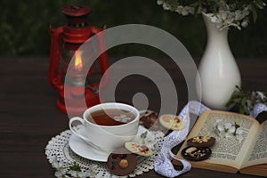 cup of tea with cherry blossom decoration on dark brown wooden background. Chokolate, bouquet, book and kerosene lamp