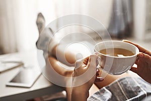 Cup of tea and chill. Woman lying on couch, holding legs on coffee table, drinking hot coffee and enjoying morning