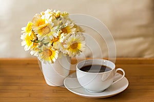 A cup of tea/coffee and a bouquet of daisies in a vase on a wooden tray on a light soft background
