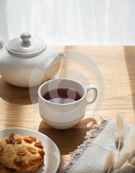 A cup of tea with homemade cookies and a white teapot on a wooden table against the background of a window with sunny morning