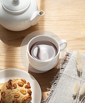 A cup of tea with homemade cookies and a white teapot on a wooden table against the background of a window with sunny morning